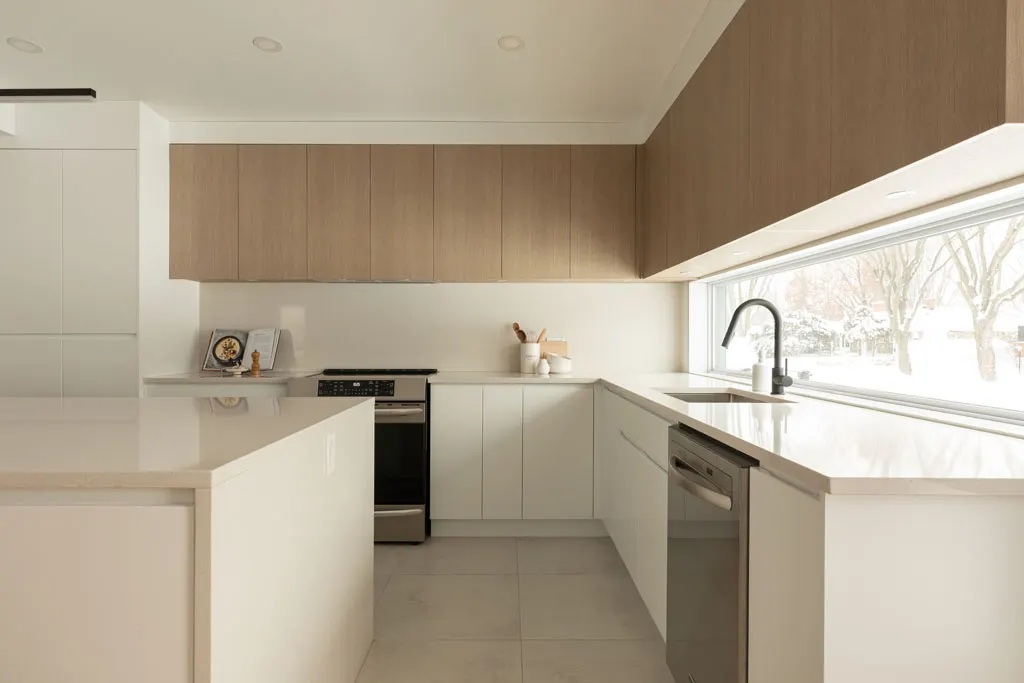 Cuisine en L vue du salon : armoires hautes de bois, cuisinière et évier sous une large fenêtre