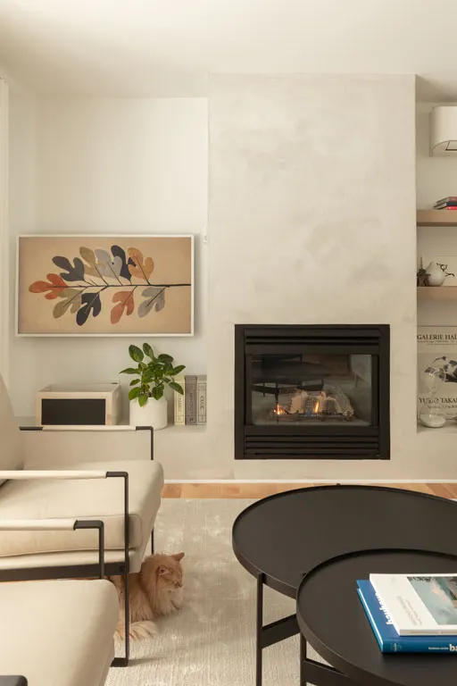Foyer au gaz allumé dans son mur d'enduit pâle, tables gigognes noires et chat roux couché au pied du fauteuil