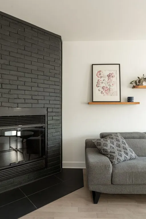 Foyer encastré dans un mur de briques peintes en noir, tablette de bois et divan gris au salon
