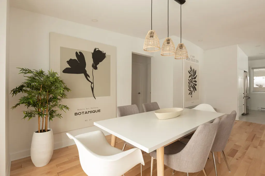 Salle à manger avec table blanche, trois suspensions en rotin et affiche botanique sur plancher de bois